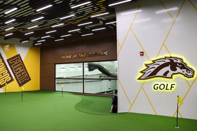 An image of Western Michigan University's indoor putting and chipping lab. The lab featured green synthetic turf, colorful wall graphics, bright lighting, and holes marked with small black and yellow flags dispersed throughout the turf.