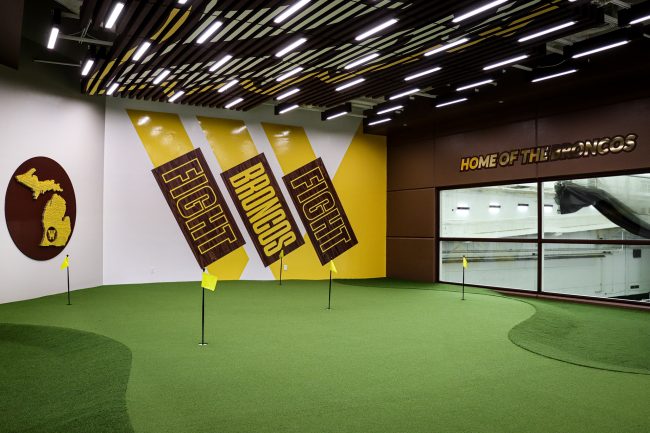 An image of Western Michigan University's indoor putting and chipping lab. The lab featured green synthetic turf, colorful wall graphics, bright lighting, and holes marked with small black and yellow flags dispersed throughout the turf.