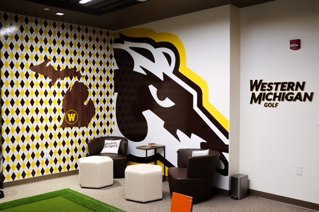 An image of Western Michigan University's indoor hitting lab seating area featuring white leather chairs topped with gold and brown pillows and colorful graphics on the wall behind.