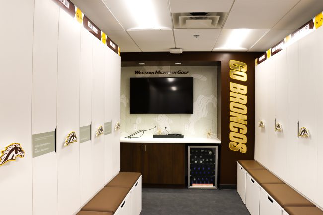 An image of Western Michigan University's women's golf locker room featuring two banks of white lockers, a television on the wall at the far end of the room as well as a mini refrigerator beneath and countertop. There are also colorful wall graphics and brown leather benches beneath lockers.