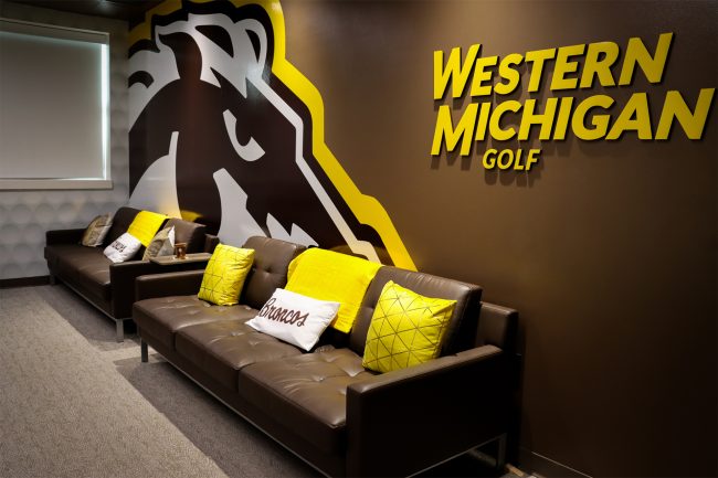 An image of Western Michigan University's indoor putting lab seating area featuring brown leather couches topped with gold and white pillows and colorful graphics on the wall behind.