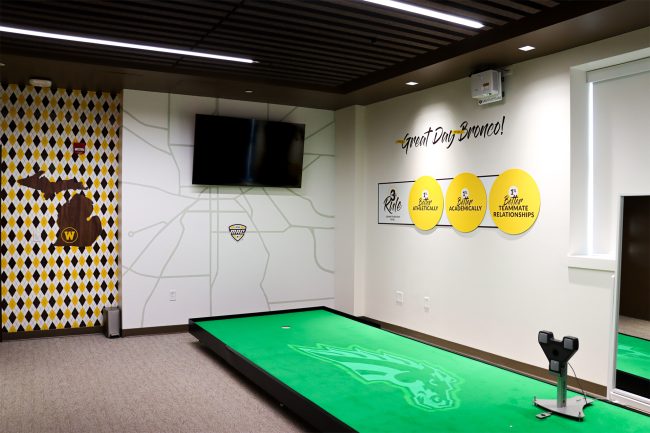 An image of Western Michigan University's indoor putting lab. The lab features an elevated green synthetic turf table that can be tilted to simulate nature. There is a screen on the wall in front of the turf to show the player how to align their aim with the hole.